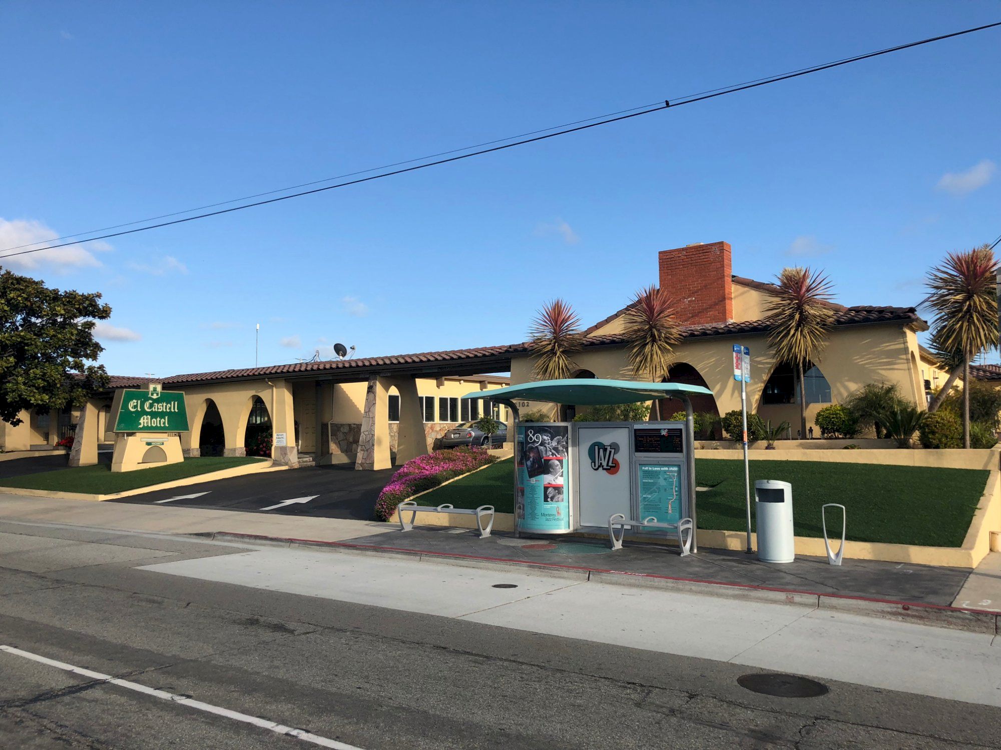 A single-story building with arches, a small parking lot, and a blue bus stop shelter on a sunny day with palm trees in the yard.