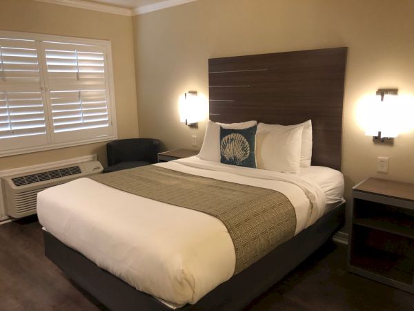A neatly made hotel room with a king bed, padded headboard, two bedside tables, wall lamps, window with blinds, and an air conditioner.