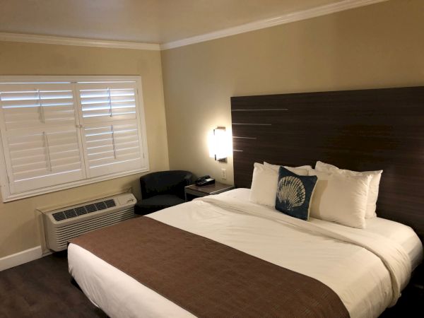 A hotel room with a king bed, white linens and brown accent throw, dark wood headboard, window with blinds, and a small seating area by the AC unit.