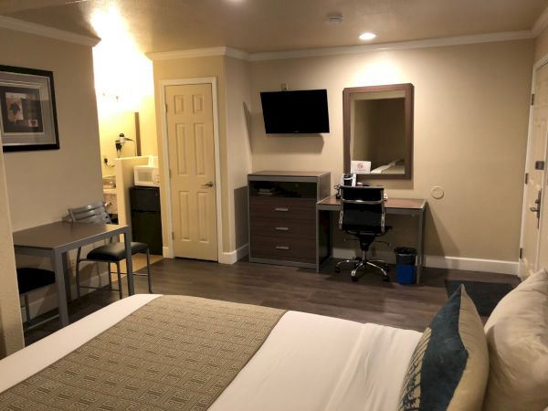 Hotel room with a bed, desk area, wall-mounted TV, dresser, mirror, and a small seating nook—neutral tones and modern furnishings.