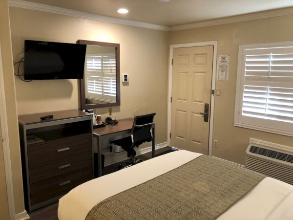 A hotel room with a bed, dresser, wall-mounted TV, desk and chair, mirror, door, blinds, and an AC unit—neat and simple, ready for guests.