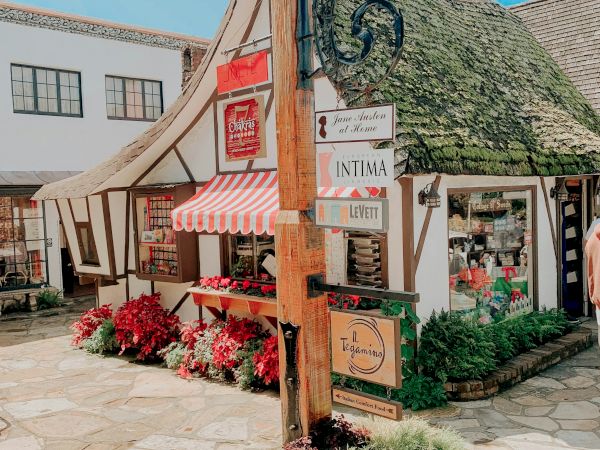 A charming European-style plaza with a thatched-roaf building, colorful storefronts, flowers, and street signs under sunny skies.