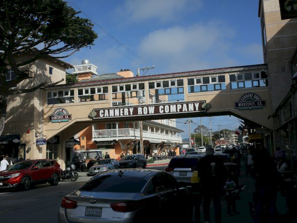 A sunlit street scene under a pedestrian bridge that reads “CANERY ROW COMPANY,” with cars, people, and buildings on either side.