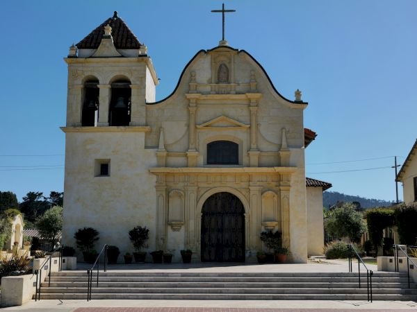 A small old church with a bell tower on the left, a large ornate entrance, steps up front, cross on top, and clear blue sky.
