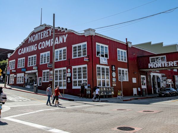 A red, two-story building labeled “Monterey Canning Company” sits on a street corner with people walking nearby under a clear sky.