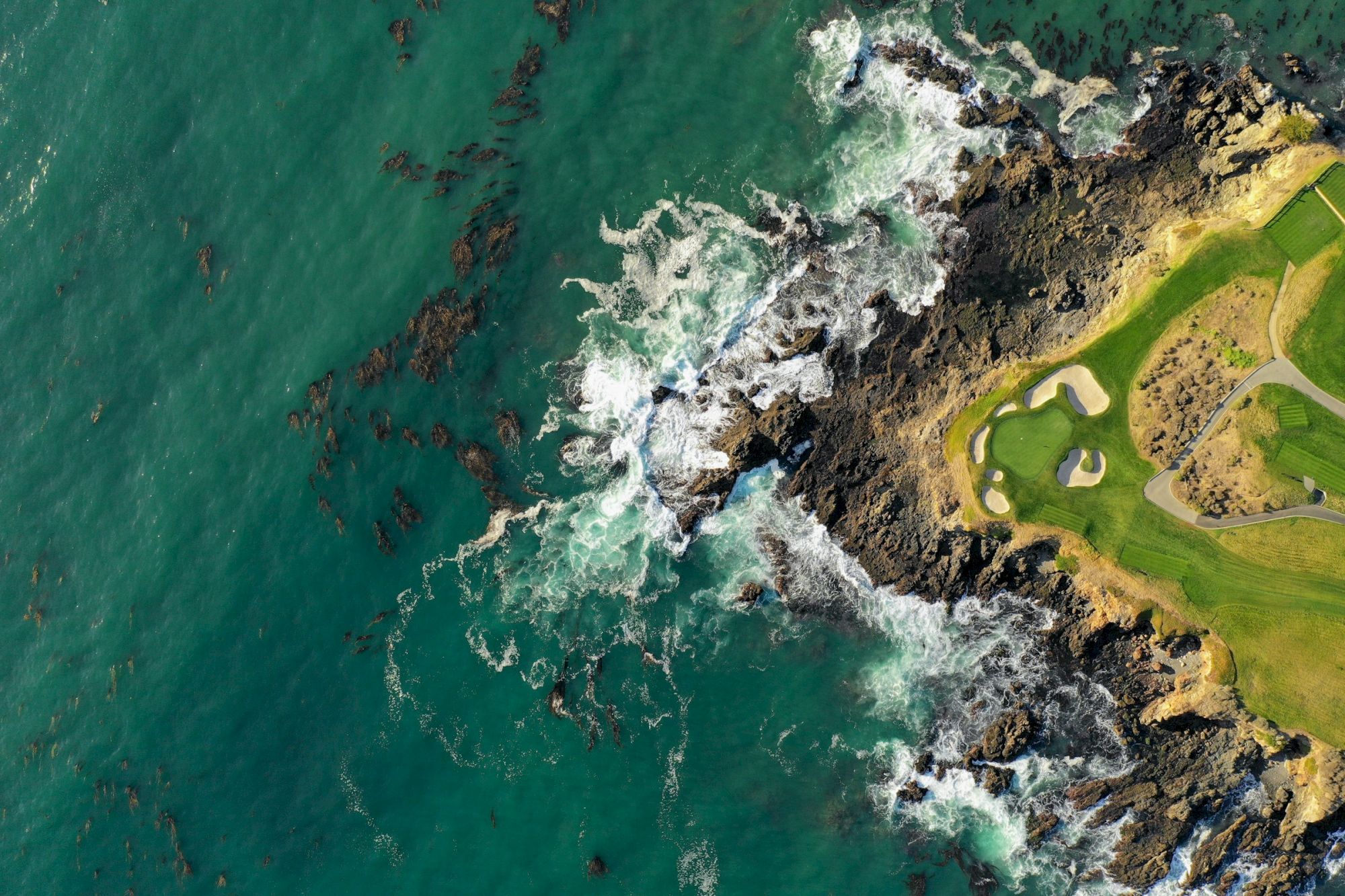 Aerial view of rugged coastline with crashing waves, rocky shoreline, and a bright green golf course area along the edge.