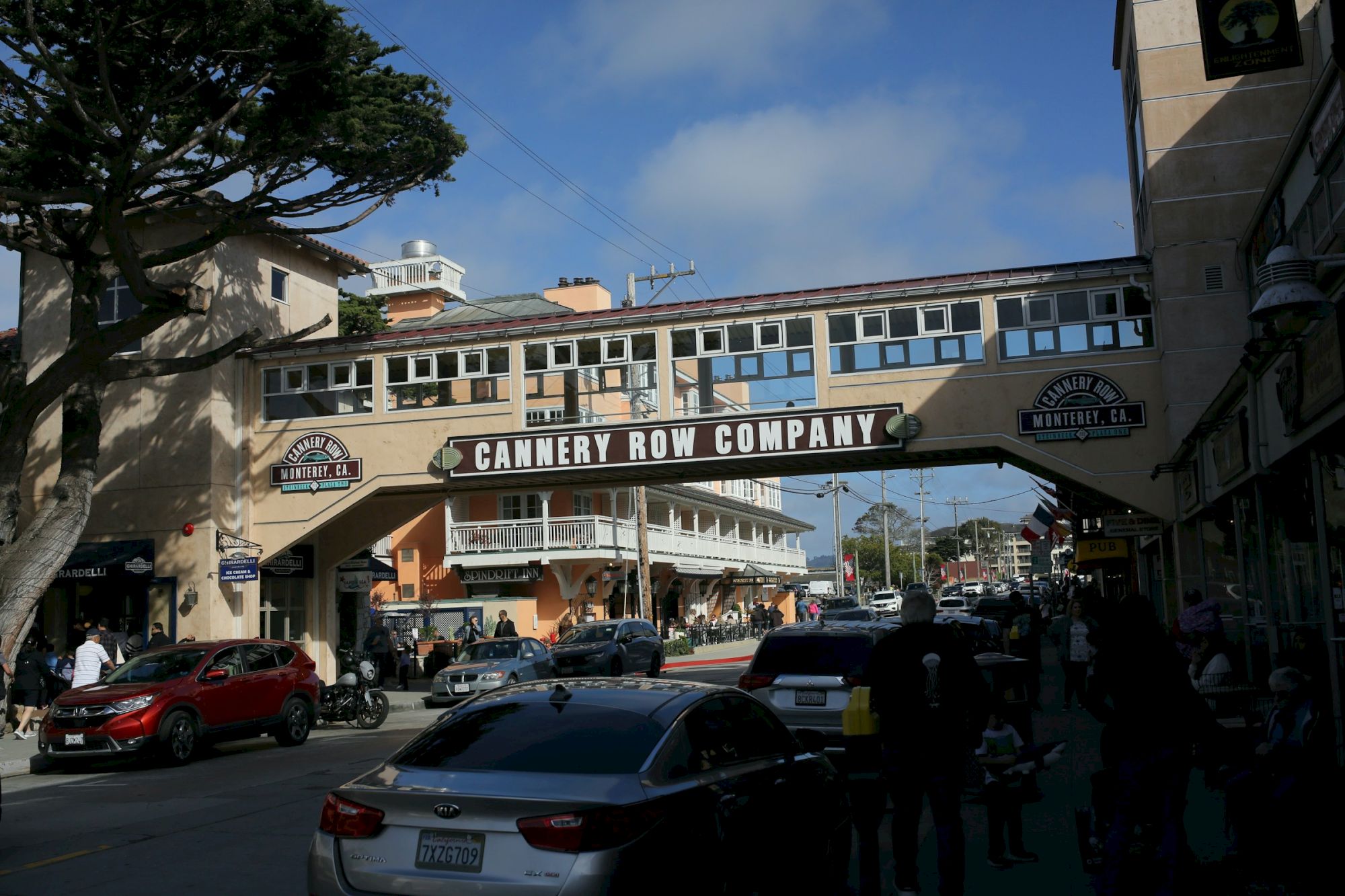 A busy street scene under a covered pedestrian bridge labeled “Cannery Row Company,” with cars, buildings, and pedestrians on a sunny day.