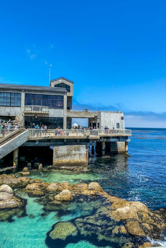 A concrete seaside building on stilts extends over turquoise water with rocks below; people on the deck enjoy the sunny coastal scene.