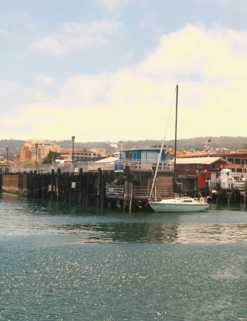 A small harbor with a wooden pier, boats docked along the quay, calm blue water, and low buildings on shore under a sunny sky.