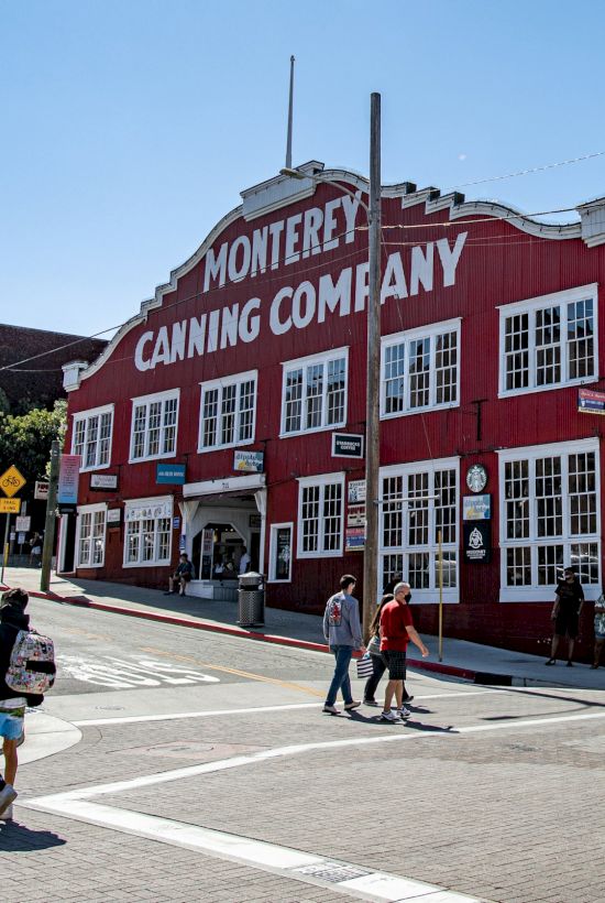 Colorful red brick building labeled “Monterey Canning Company” at a sunny street corner with pedestrians crossing.