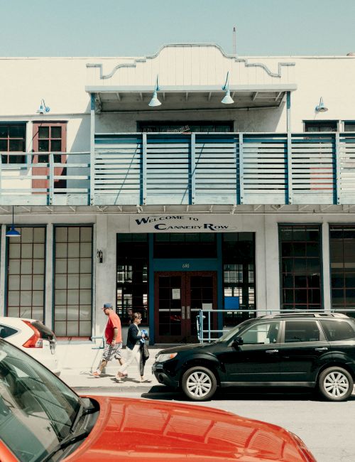 A small town storefront with a pale building, a striped awning, people walking on the sidewalk, and several parked cars in the street. End.