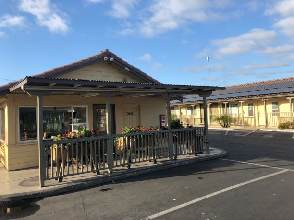 A small, single-story building with a covered walkway and a railing, likely part of a shopping or service center, under a sunny, partly cloudy sky.