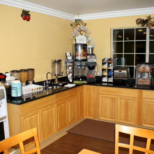 A cozy kitchen setup with yellow walls, wooden cabinets, a long counter, coffee gear, a water cooler, and a small dining table nearby.