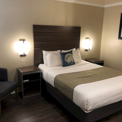 A hotel room with a queen bed, two nightstands, wall lamps, a chair, and framed art on the wall. End.