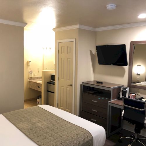 A tidy hotel room with a bed, dresser, wall-mounted TV, work desk, mirror, and a small sitting area near a doorway, neutral tones throughout.