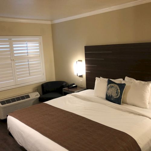 A hotel-style bedroom with a large bed, brown and white bedding, a headboard, window with blinds, an air conditioner unit, a chair, and bedside lights.