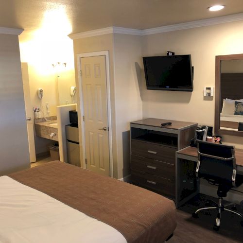A hotel room with a bed, desk and chair, dresser, wall-mounted TV, mirror, entry to a bathroom, and neutral decor, cozy and neat.