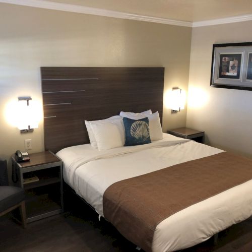 A hotel room with a king bed, brown headboard, two bedside tables, wall lamps, a framed picture, and a chair in the corner.