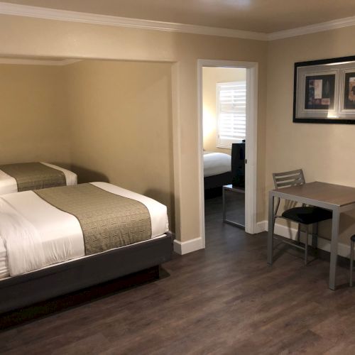 A hotel-style room with two beds in a alcove, a small desk and chairs, and a doorway to a separate room; neutral decor.