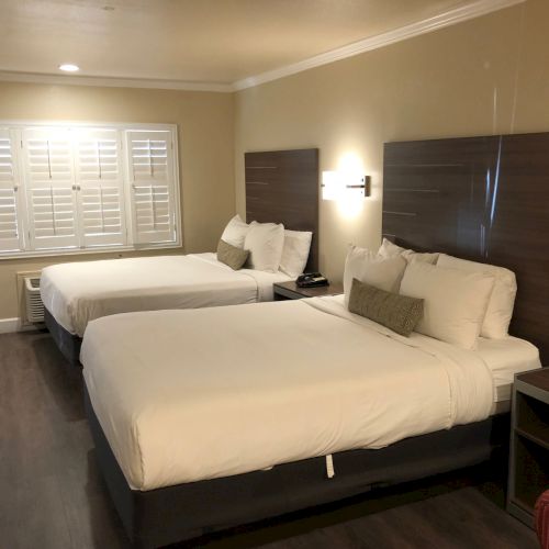 A hotel room with two large beds, dark wooden headboards, nightstands, a window with blinds, and cozy lamps by the beds.
