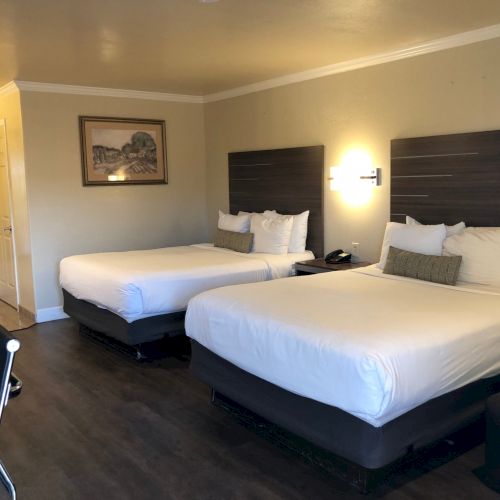 A hotel room with two queen beds, dark wood flooring, a wall-mounted light, a seating chair, and a doorway with a mirror in the corner.