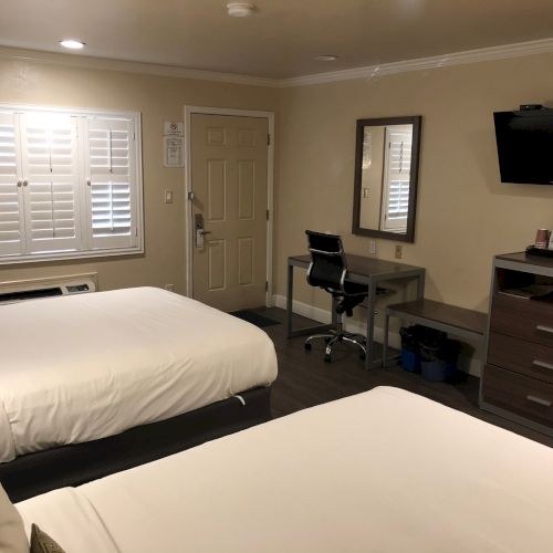 Two neatly made beds in a hotel room, with a desk, chair, wall-mounted TV, dresser, mirror, and a window with blinds. End of sentence.