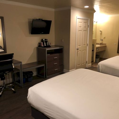 Two double beds in a hotel room with a desk, chair, wall mirror, small TV, dresser, and a private bathroom area in the background.