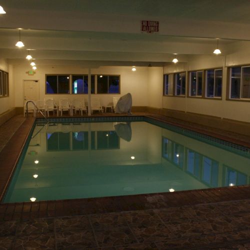 An indoor swimming pool area with a rectangular pool, tiled deck, ceiling lights, and windows along the walls, empty and quiet. ending sentence.