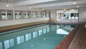 Indoor swimming pool area with clear water, tiled deck, pendant lights, and a lounge/rec room visible in the background.