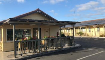 A small single-story building with a wraparound porch and railing, potted plants, and a parking lot in front under a blue sky.