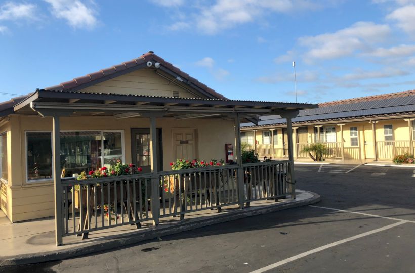 A small single-story building with a wraparound porch and railing, potted plants, and a parking lot in front under a blue sky.