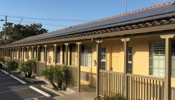 A single-story beige motel/row of rooms with porches, railings, small shrubs, and a parking lot in front, sunny day.