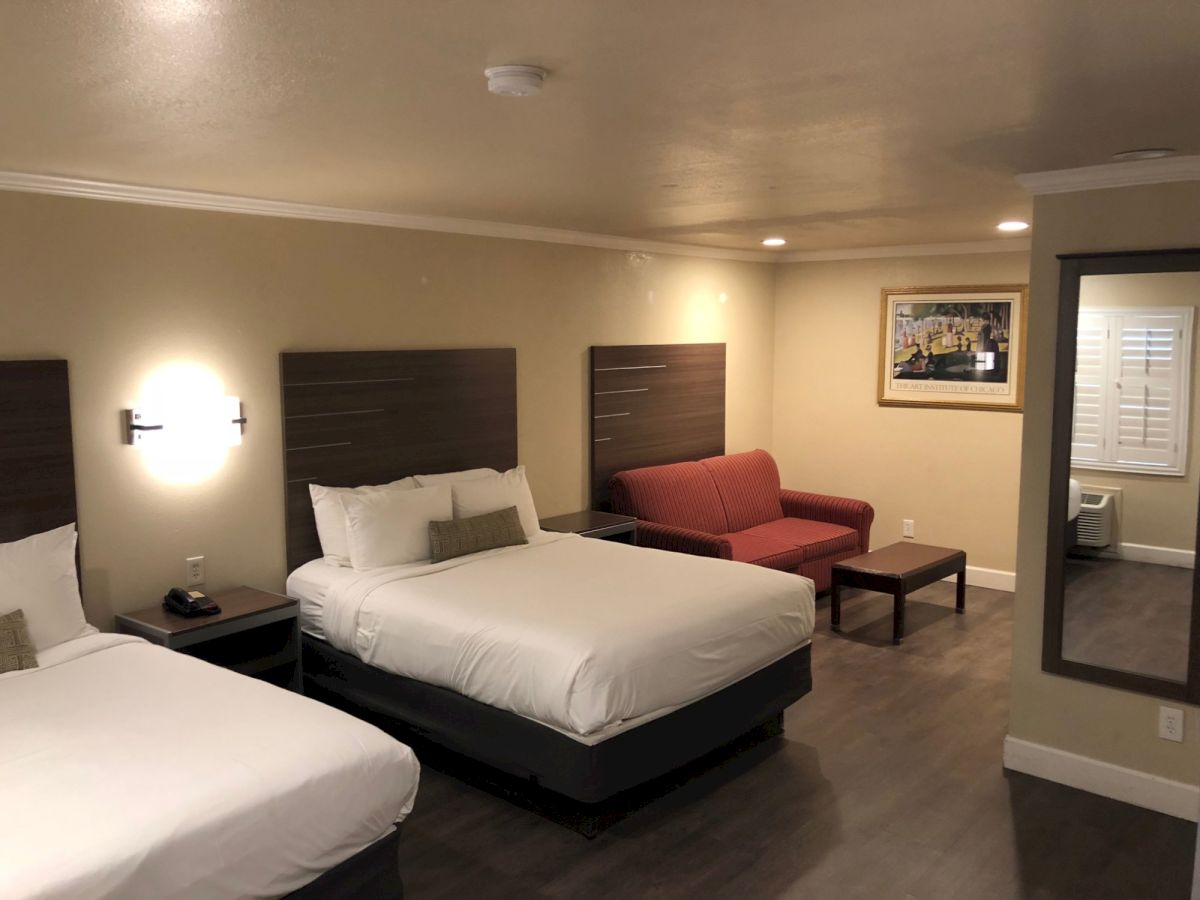 Three beds in a hotel room with a red sofa, coffee table, framed art, and a bathroom visible through an open doorway.