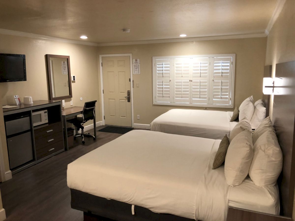 Hotel room with two double beds, desk, chair, mini-fridge, and TV; neutral decor, window with shutters, soft lighting.