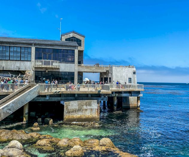 A seaside pier with a concrete building on stilts, people on the deck, clear blue sky, and turquoise water with rocks along the shore.