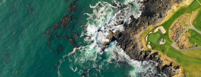 Coastal cliff with rocks meeting turquoise waves, white foam, and a grassy shoreline forming a rugged shoreline scene.